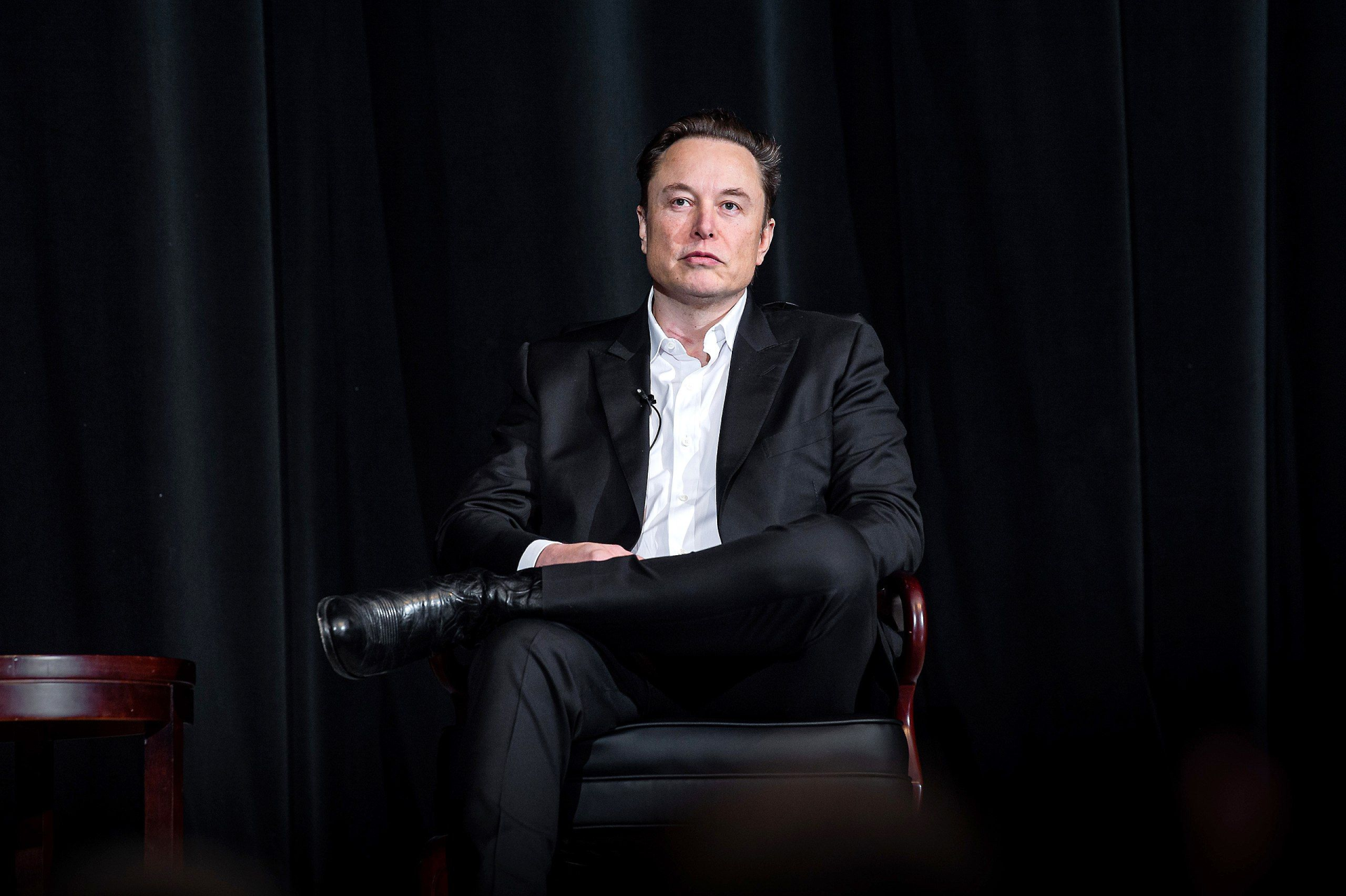 A photo of Elon Musk sitting on the chair against the black background. 