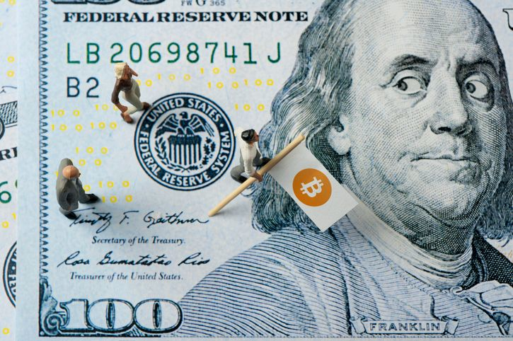 A stock photo of a surprised one hundred dollar banknote being walked over by a group of three people with Bitcoin flag.