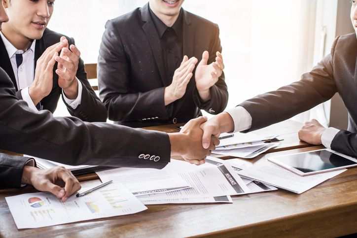 A stock photo of two businessmen shaking hands while two others cheer.