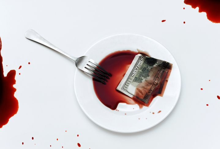 A hundred-dollar bill lies in a white plate next to blood-like soy sauce with the fork on tablecloth. 