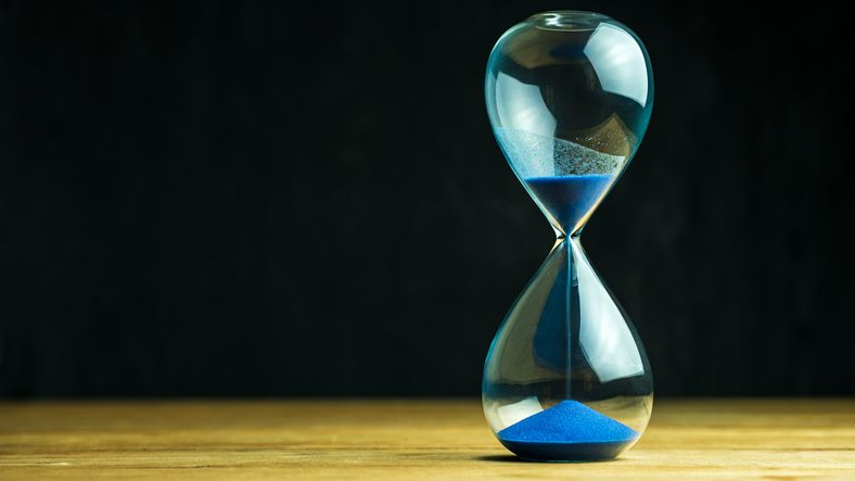 A stock photo of an hourglass with blue sand against the black background.