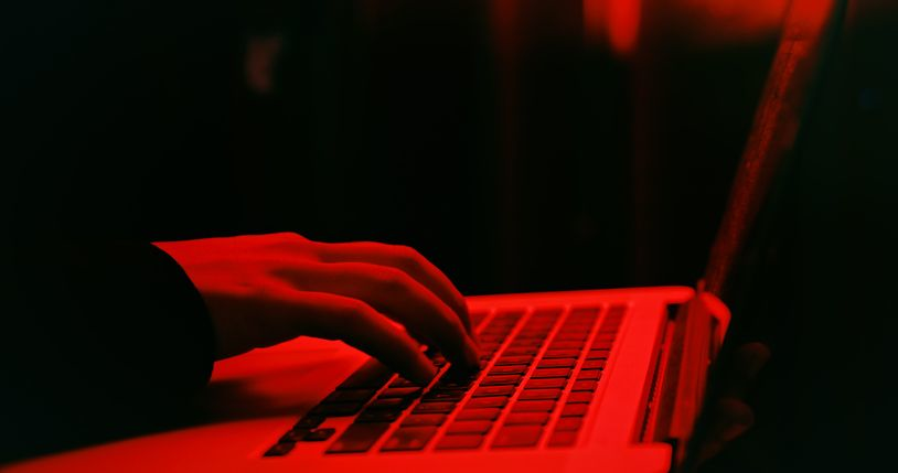 A stock photo of a hand over the laptop in neon red light, defi hacker concept. 