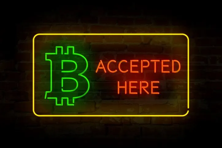 A stock photo of a neon Bitcoin logo and "accepted here" inscription. 