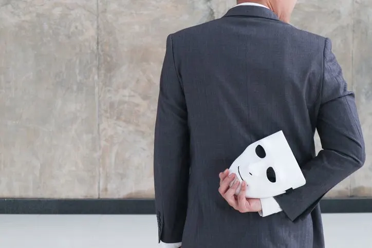 A stock image of a man hiding a white theatre mask behind his back.