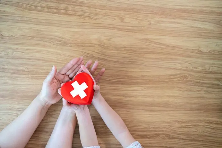 Red Heart with cross in hands of the child and mother on a white wooden 