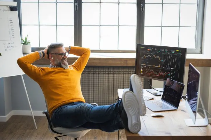 Happy trader in yellow jumper looking at his trading stations with his feet on the desk