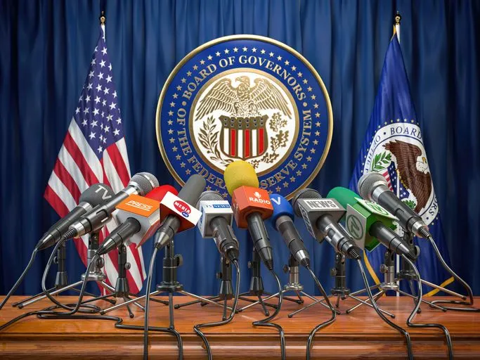 A stock photo of tv and radio microphones directed at the empty seat under the Fed logo. 