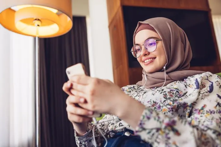 A stock photo of a Middle eastern woman in hijab using her smartphone with headphones. 