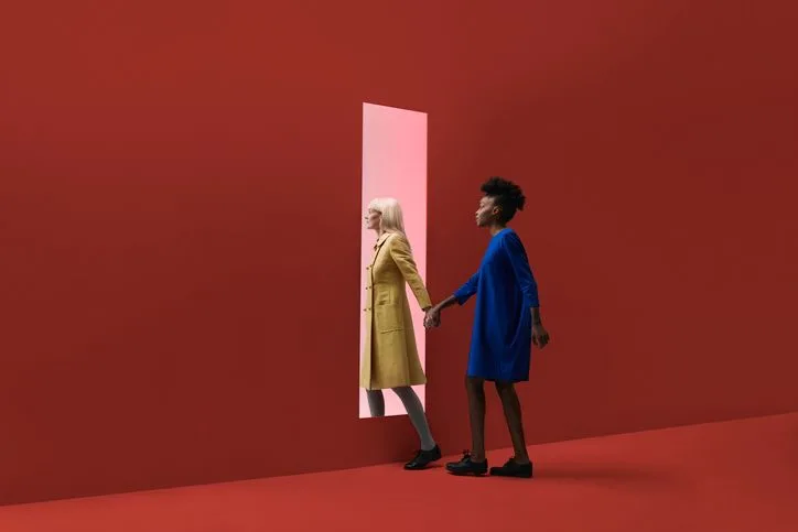 A stock photo of two women holding hands while going through a rectangular portal in the red wall. 