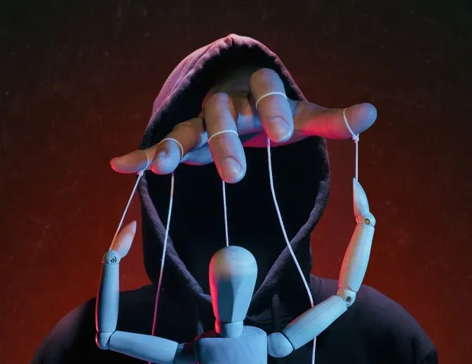 A stock photo of a man in a hoodie pulling strings of a puppet. 