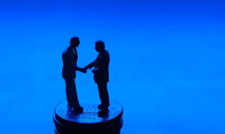 A stock photo of two businessmen shaking hands on the stack of coins against blue background. 