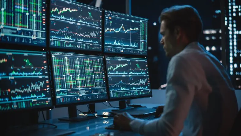Crypto trader analysing financial charts on multiple monitors in a dark room. 